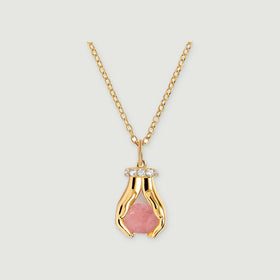 A yellow gold vermeil necklace with a raw pink quartz pendant framed by two sculpted hands and clear stones on a pale gray surface.