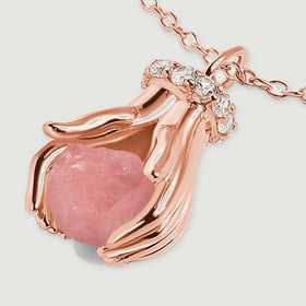 An angled close-up shows a pink quartz nugget between rose gold sculpted hands beneath a row of clear stones.