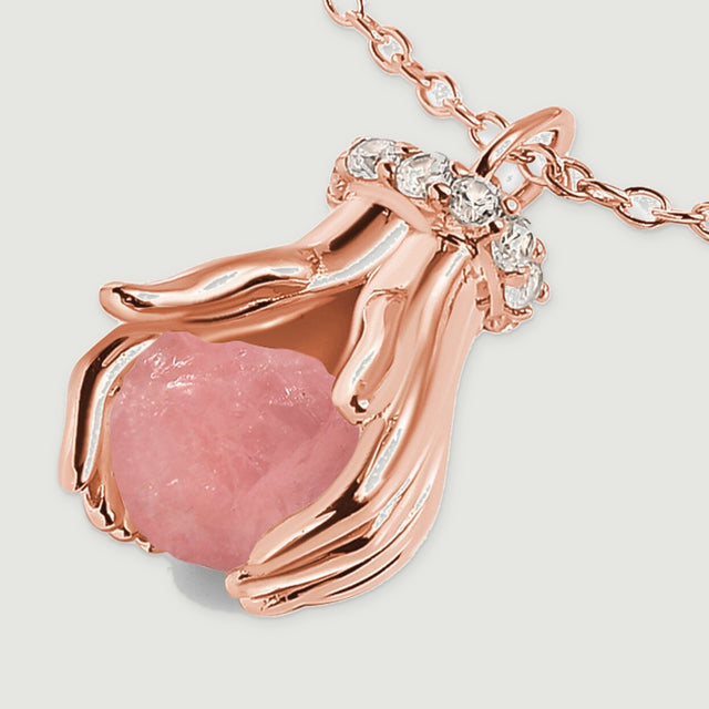 An angled close-up shows a pink quartz nugget between rose gold sculpted hands beneath a row of clear stones.