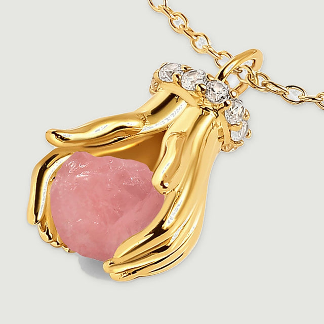 A yellow gold pendant rests diagonally with its textured pink stone, curved fingers, and circular chain links in view.