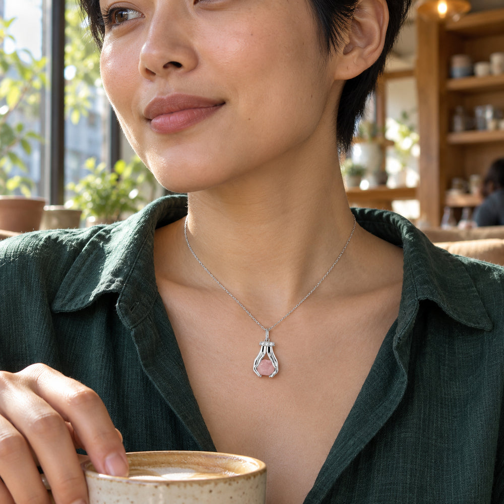 A woman with short black hair wears a silver pink-quartz necklace above a dark green shirt beside a cafe window.
