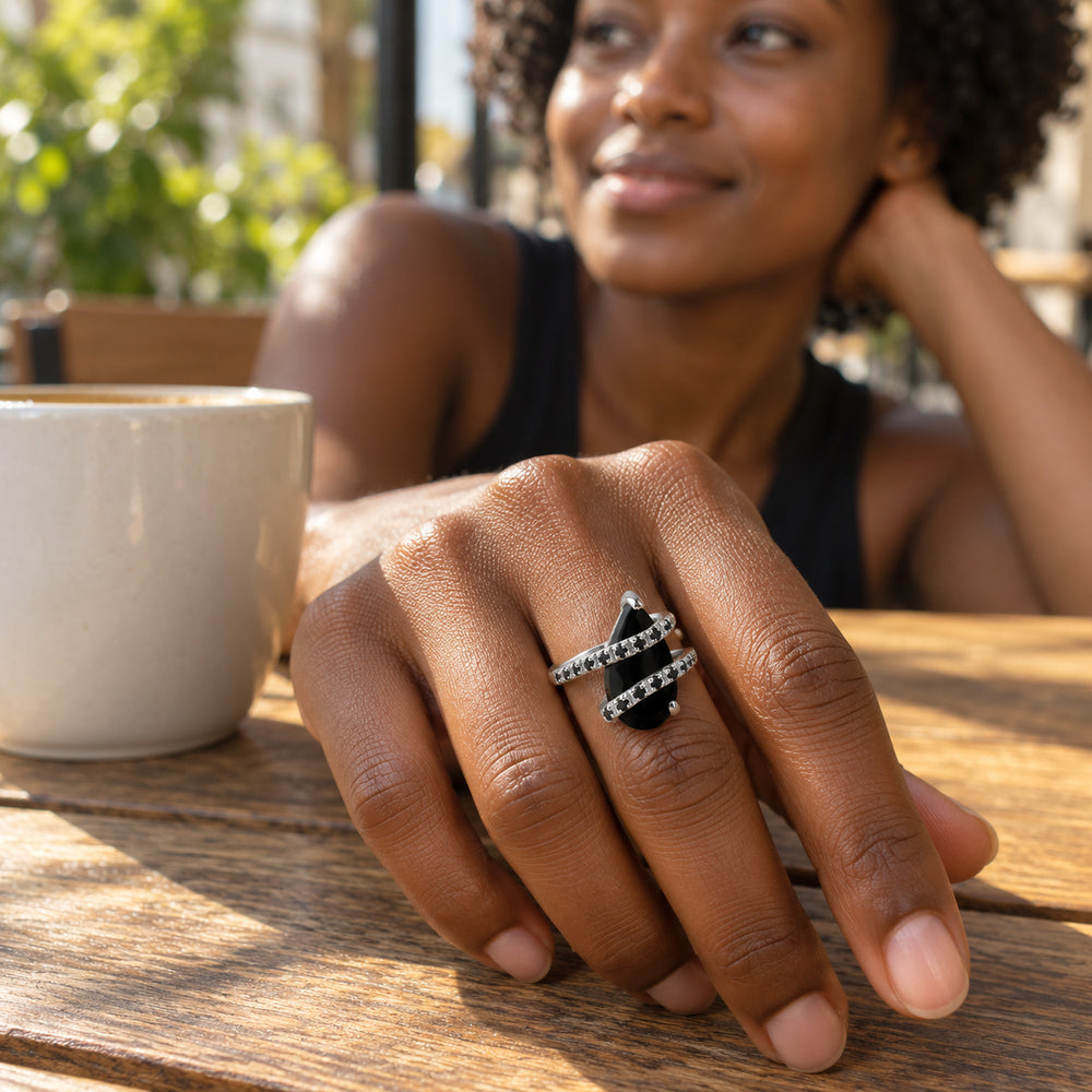 A silver ring with a black pear-shaped stone is worn on a hand resting beside a ceramic cup on a wooden cafe table.