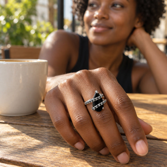 A silver ring with a black pear-shaped stone is worn on a hand resting beside a ceramic cup on a wooden cafe table.