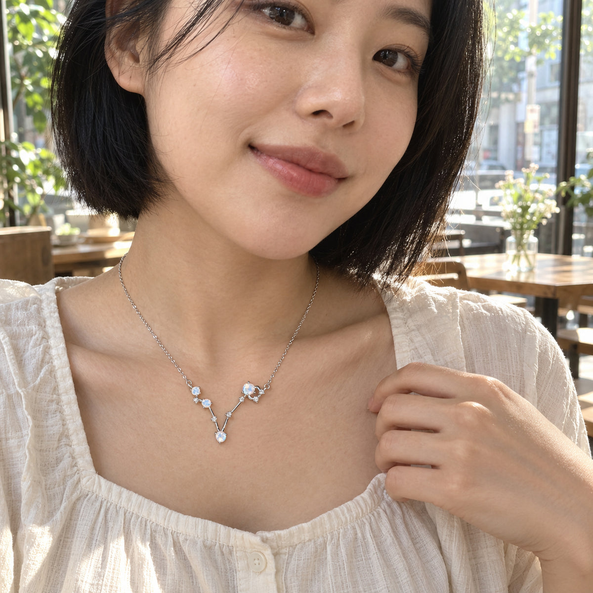 A woman with a short black bob wears a silver necklace with an open V-shaped pendant above a cream blouse in a sunlit cafe.