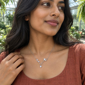 A woman with long dark waves wears a silver necklace over a rust-colored linen top among plants in a glass conservatory.