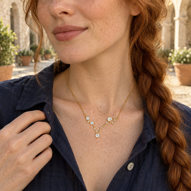 A freckled woman with a copper braid wears a yellow gold necklace above a navy shirt in a stone courtyard.