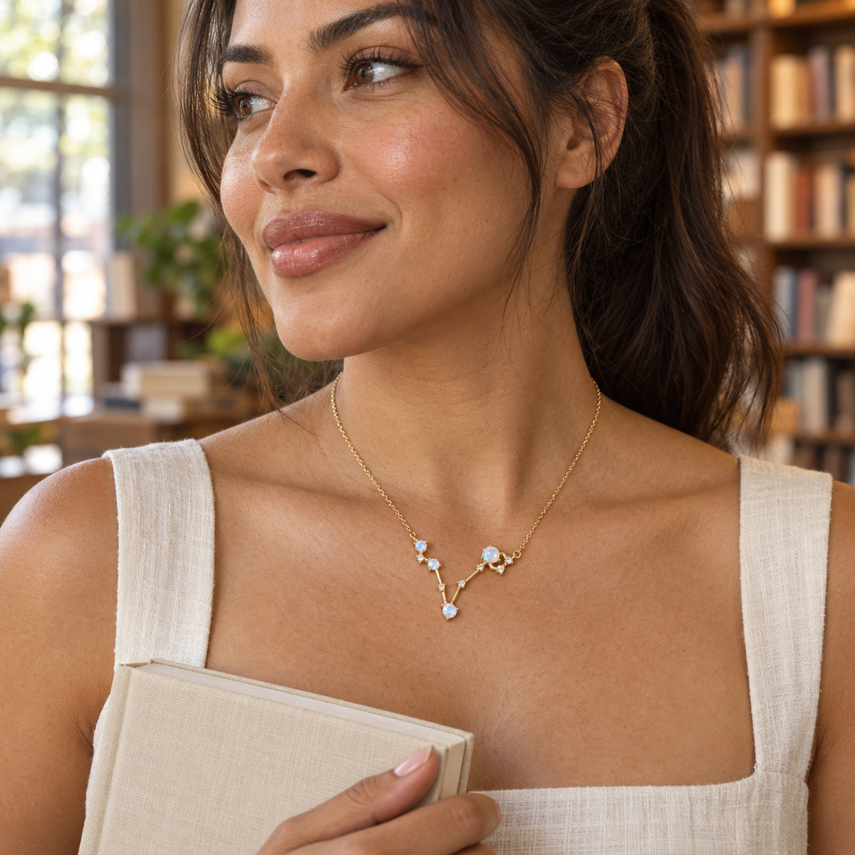 A woman with a dark ponytail wears a yellow gold necklace and an ivory dress while holding a plain book beside wooden bookshelves.