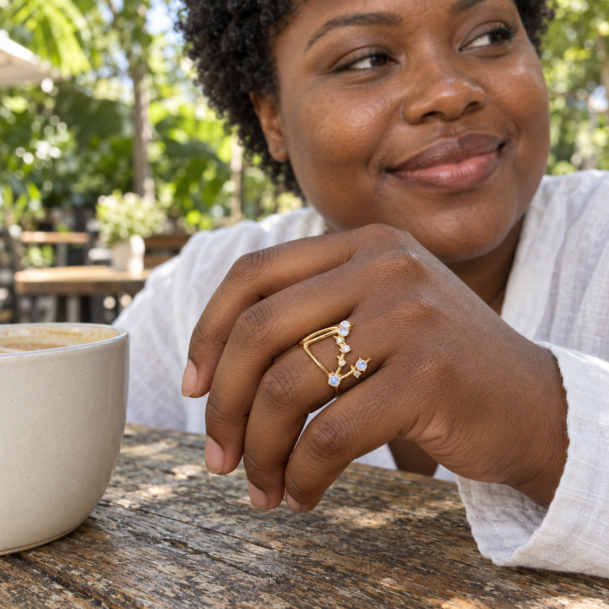 A woman with deep brown skin wears the yellow gold ring while resting her hand beside a white coffee cup at an outdoor cafe.