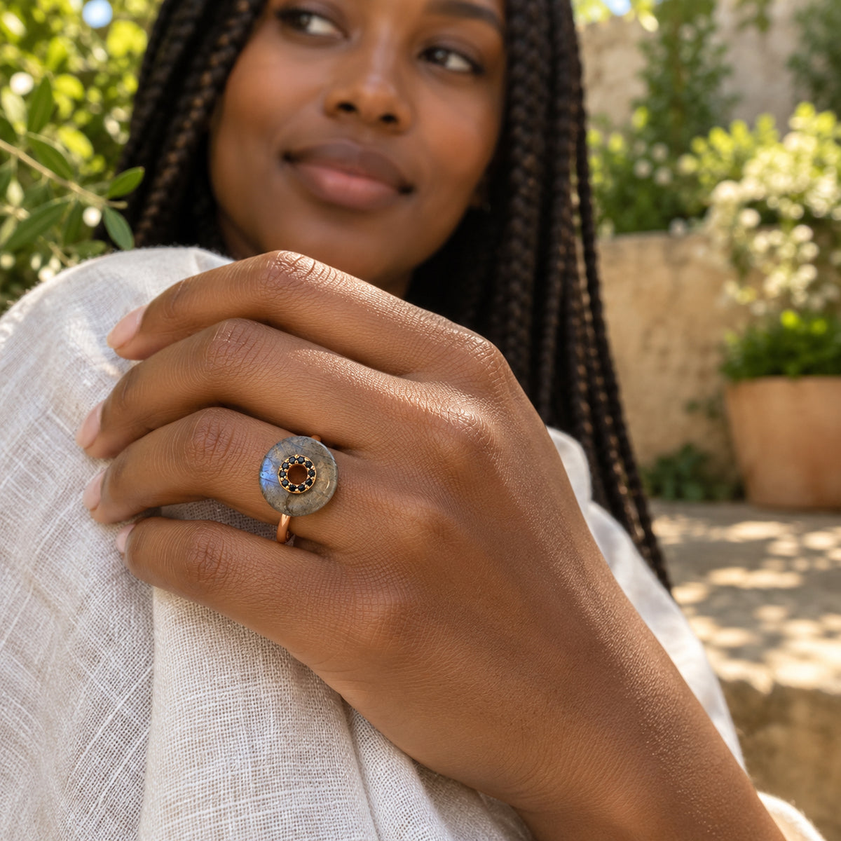 A hand wearing a rose gold vermeil labradorite ring rests against pale linen with a sunlit garden in the background.