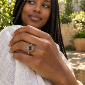 A hand wearing a rose gold vermeil labradorite ring rests against pale linen with a sunlit garden in the background.