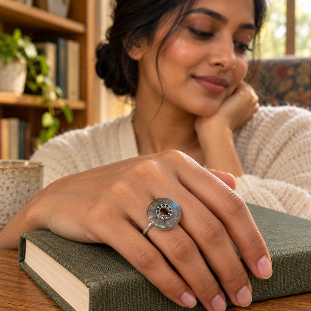 A sterling silver labradorite ring is shown on a hand resting on a closed green clothbound book near wooden shelves.