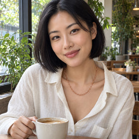 A woman in a white linen shirt wears a rose gold tone chain while holding a coffee cup in a leafy cafe.
