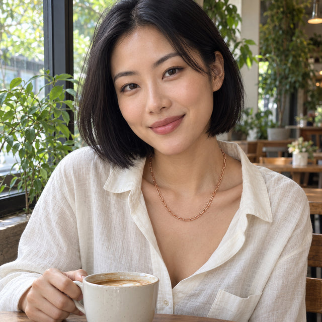 A woman in a white linen shirt wears a rose gold tone chain while holding a coffee cup in a leafy cafe.