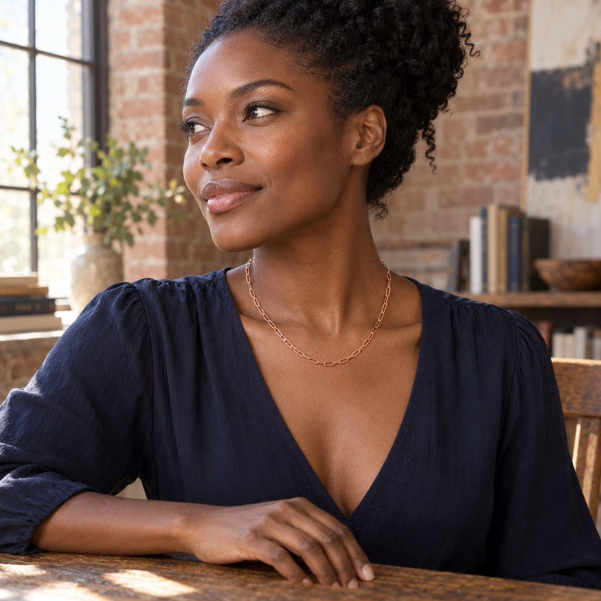 A woman in a navy blouse wears a rose gold tone necklace at a wooden table in a room with brick walls.