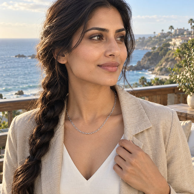 A woman with a dark braid wears a silver tone necklace and a linen jacket on a balcony overlooking the sea.