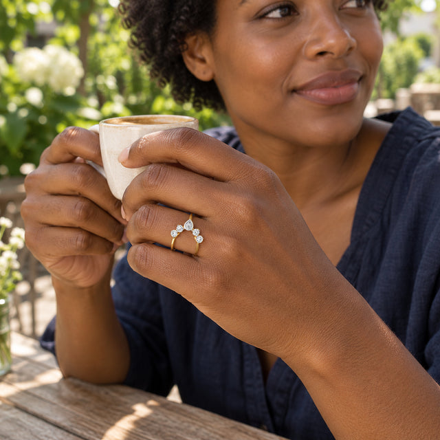 A yellow gold diamond ring is worn on a hand holding a ceramic cup in a leafy outdoor cafe.