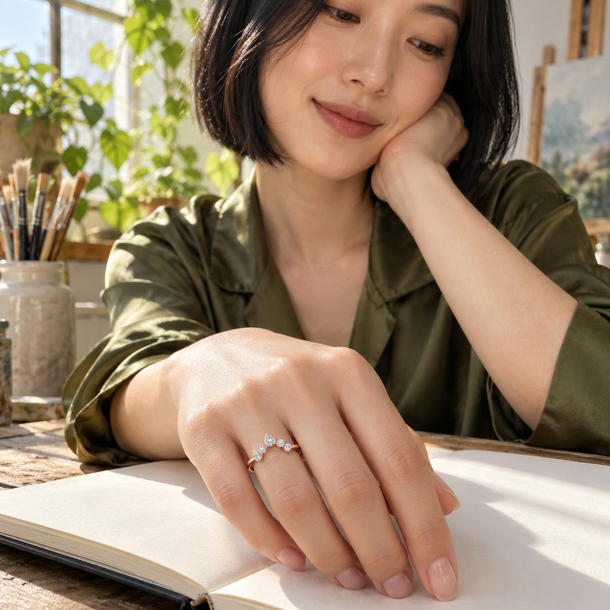 A rose gold diamond ring is shown on a hand resting over a blank sketchbook in a sunlit studio.