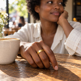 A woman wears a polished yellow gold band beside a ceramic coffee cup at a sunlit cafe table.