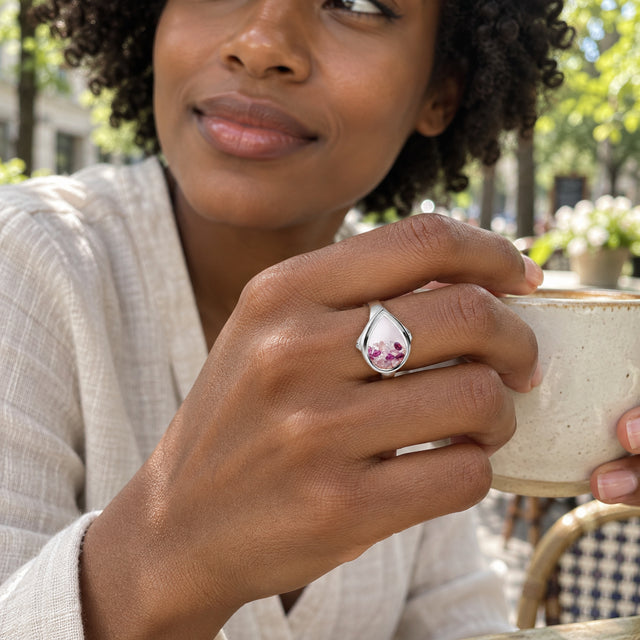A sterling silver teardrop ring with pink and raspberry stones is worn on a hand holding a ceramic cup at an outdoor cafe.