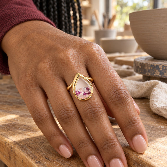 A yellow gold vermeil ring with a clear pear chamber is shown on a hand resting on a wooden pottery bench.