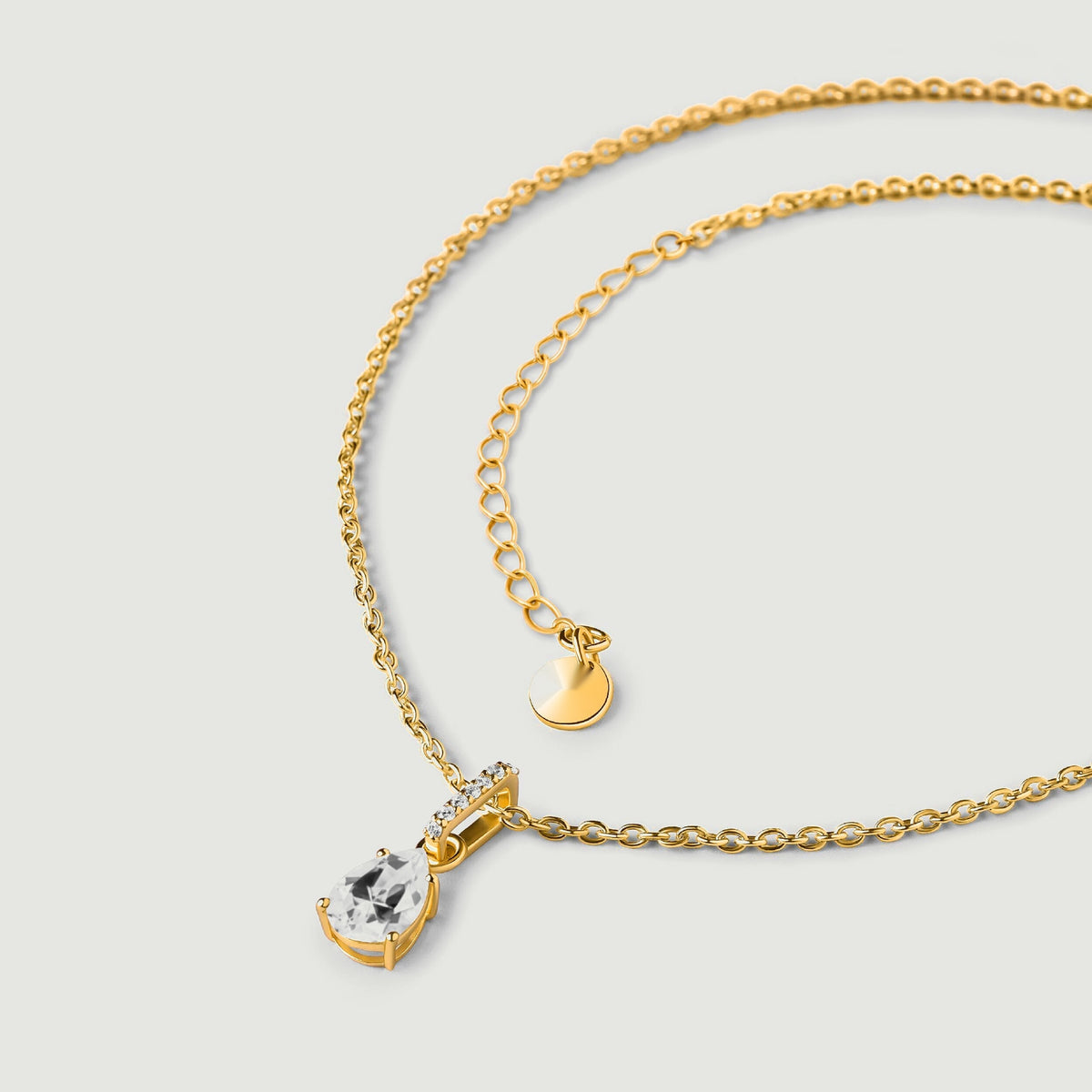 A yellow gold vermeil necklace is arranged on a clean light gray surface with the clear pendant at the lower left.