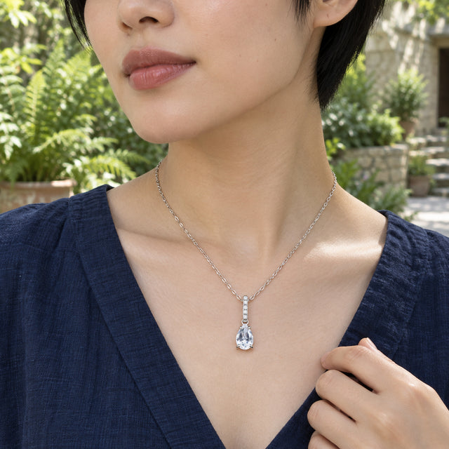 A woman in a navy blouse wears the sterling silver necklace with a clear pear-shaped pendant in a leafy courtyard.