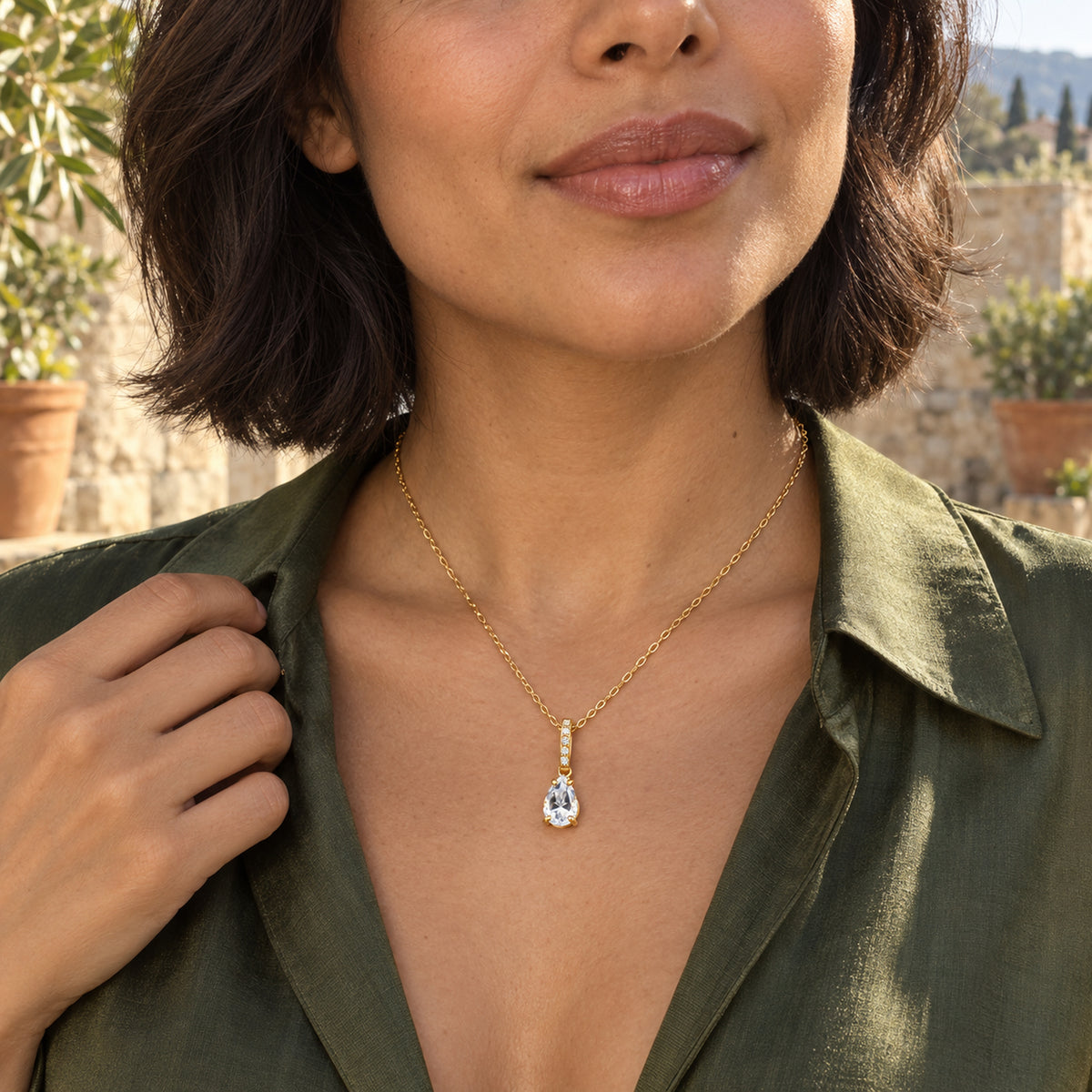 A woman with a dark bob wears the yellow gold vermeil necklace above an olive shirt on a sunlit stone terrace.