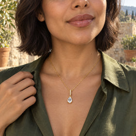 A woman with a dark bob wears the yellow gold vermeil necklace above an olive shirt on a sunlit stone terrace.