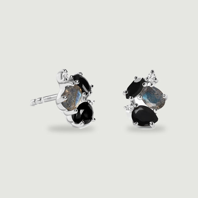 A pair of sterling silver cluster studs shows black faceted stones, blue grey labradorite and small clear accents on a light grey surface.