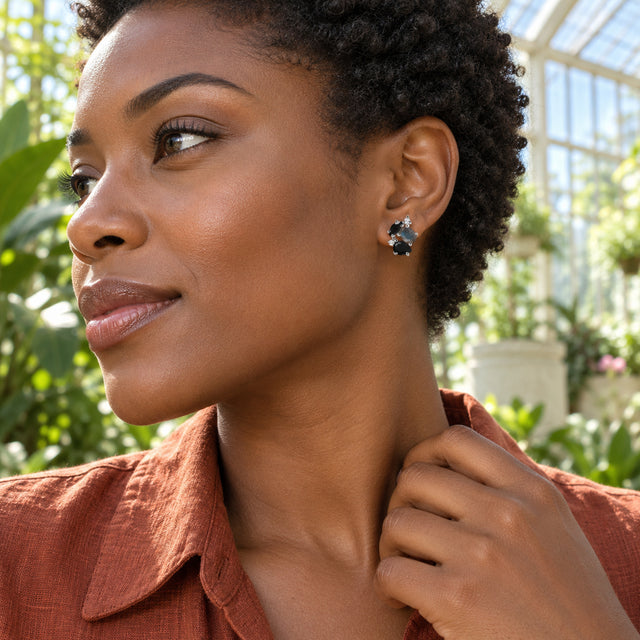 A woman with short coiled hair wears a silver cluster stud beside sunlit conservatory plants.