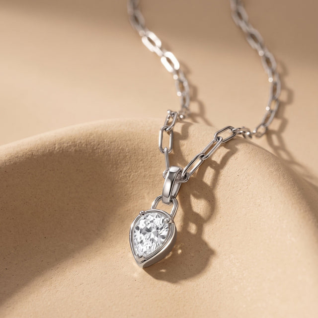 A silver-tone link necklace lies across a curved beige ceramic surface with its clear pear-shaped pendant in the foreground.