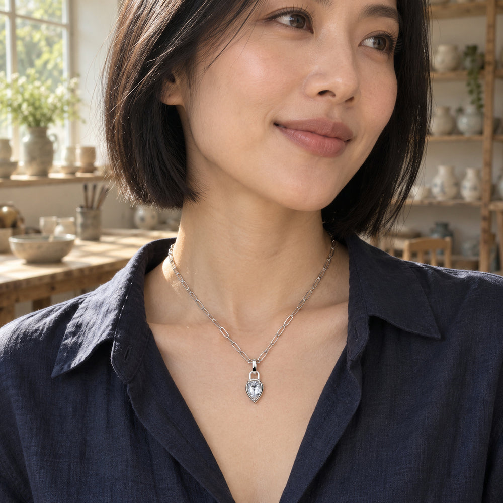 A woman with a black bob wears a silver-tone pear-shaped pendant necklace with a navy shirt in a sunlit ceramics studio.