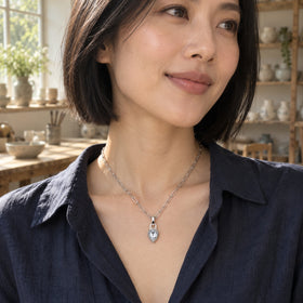 A woman with a black bob wears a silver-tone pear-shaped pendant necklace with a navy shirt in a sunlit ceramics studio.
