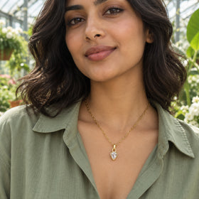 A woman with wavy dark hair wears a yellow-gold-tone necklace with a clear teardrop pendant inside a greenhouse.
