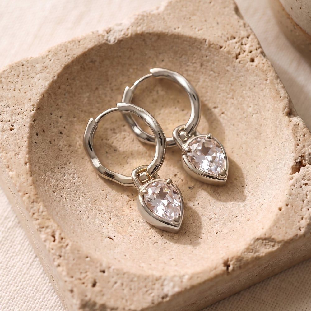Two silver-toned hoops with clear pear-shaped drops lie diagonally on a textured stone tray.