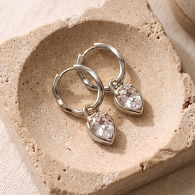 Two silver-toned hoops with clear pear-shaped drops lie diagonally on a textured stone tray.