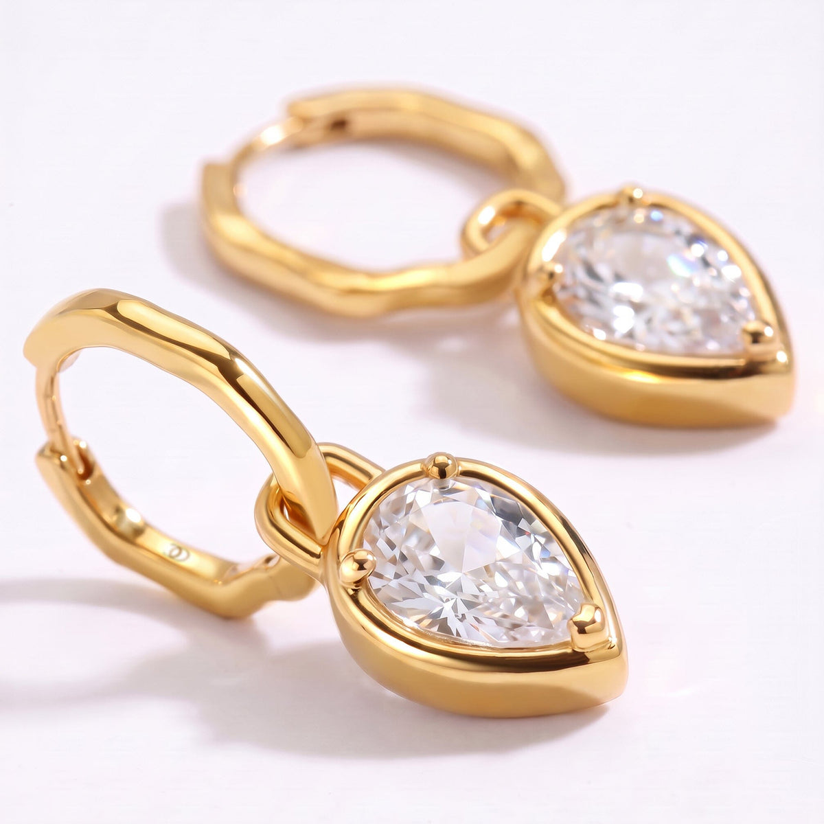 The foreground earring shows a pear-shaped clear stone in a yellow-gold setting with a second hoop behind it.