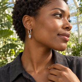 A woman with short natural curls wears a silver-toned hoop with a clear pear-shaped drop among sunlit greenhouse plants.