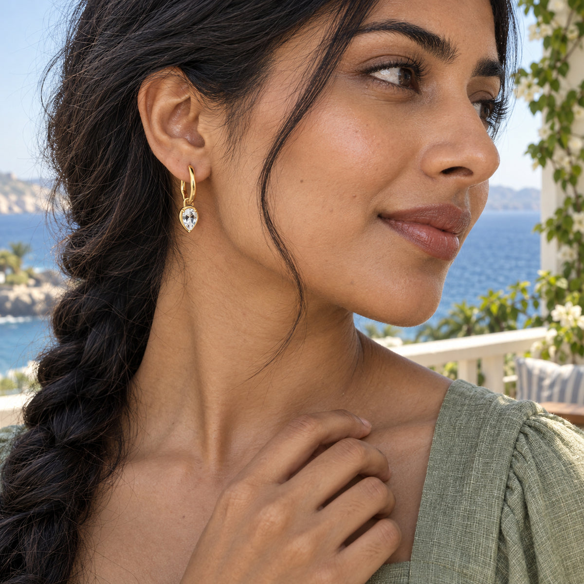 A woman with dark braided hair wears a yellow-gold hoop with a faceted clear drop on a terrace overlooking the sea.