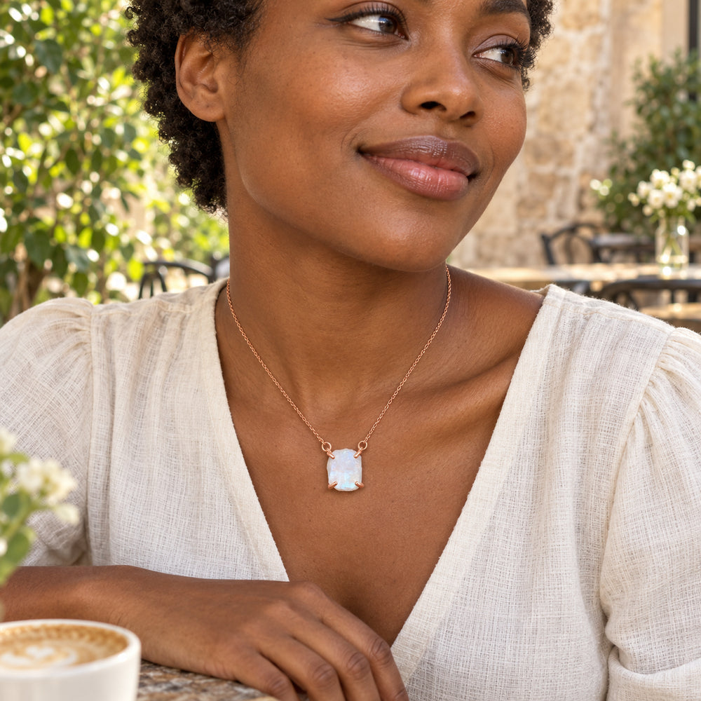 A woman in a cream linen blouse wears the rose gold moonstone necklace at a leafy outdoor cafe.