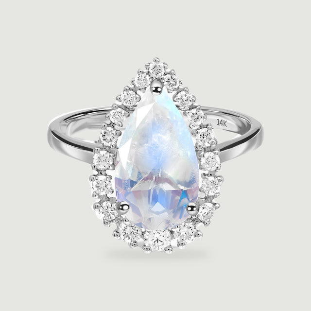 A white gold ring with a pear-shaped moonstone and a halo of round diamonds is centered on a light gray surface.