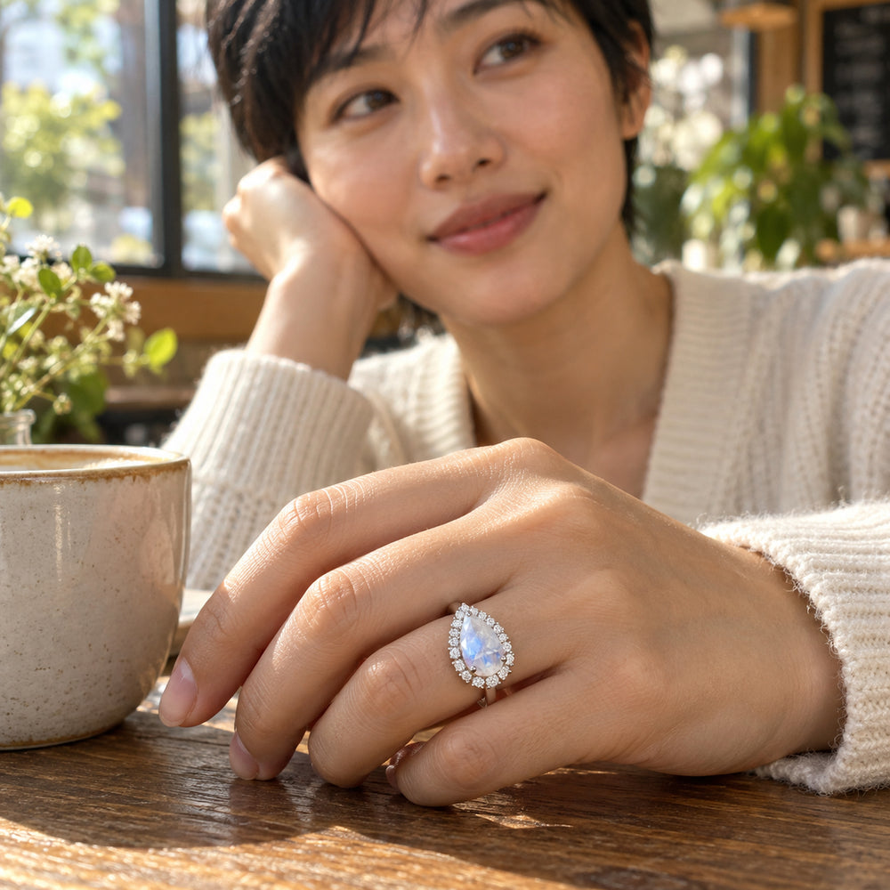A woman wears the pear-shaped moonstone halo ring with her hand resting beside a ceramic cup on a wooden cafe table.