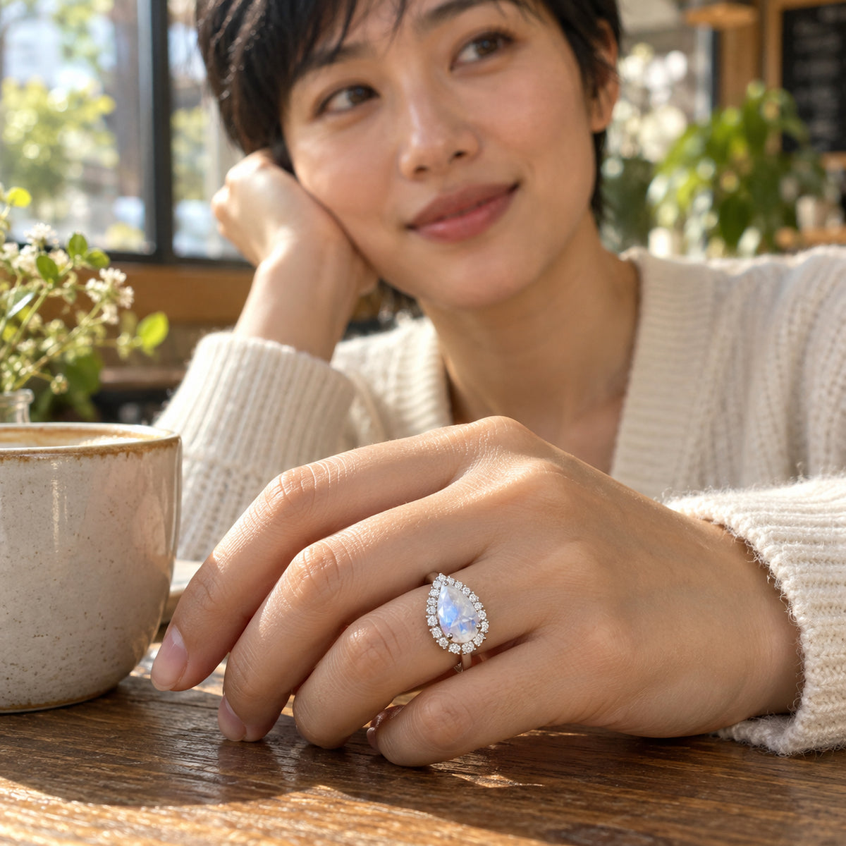 A woman wears the pear-shaped moonstone halo ring with her hand resting beside a ceramic cup on a wooden cafe table.