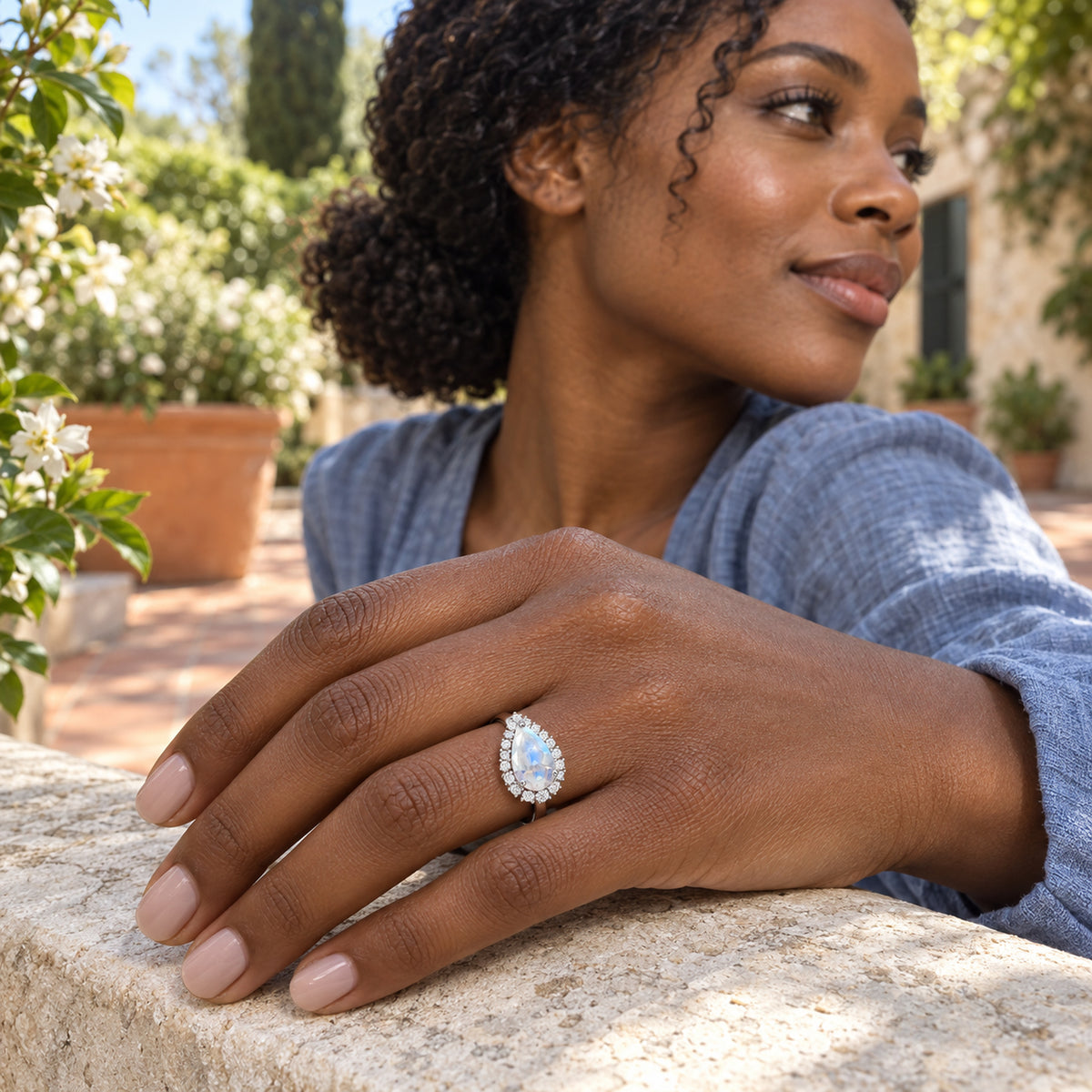 The moonstone and diamond halo ring is worn on a hand resting on a pale stone ledge in a leafy courtyard.