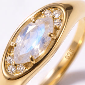 The faceted moonstone reflects pale blue light inside a polished yellow gold setting with small clear stones.