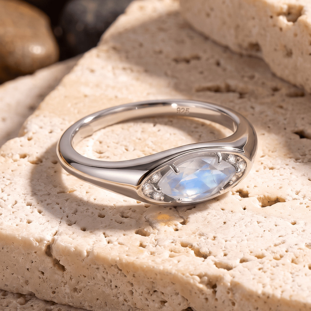 A silver moonstone ring rests at an angle on a textured cream stone surface.