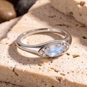 A silver moonstone ring rests at an angle on a textured cream stone surface.