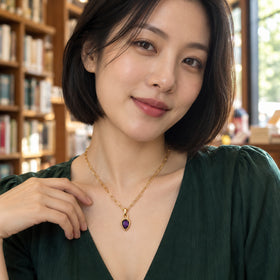 A woman in a dark green blouse wears a gold-colored link necklace with a purple pendant in a sunlit bookshop.