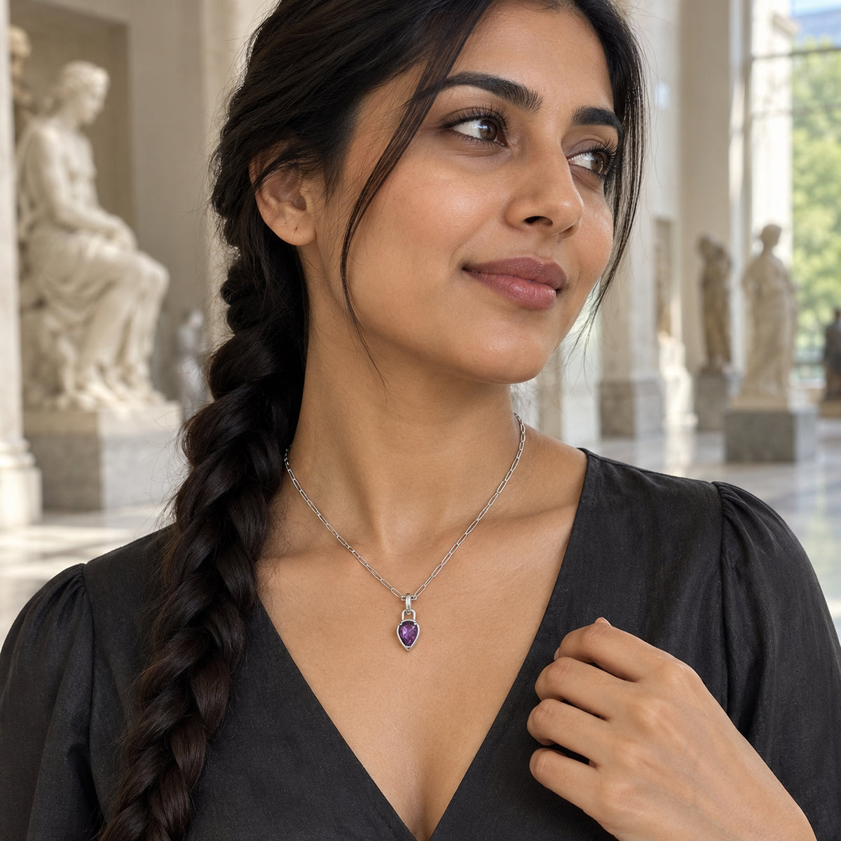 A woman with a long braid wears a silver-colored amethyst necklace with a charcoal blouse in a sculpture gallery.