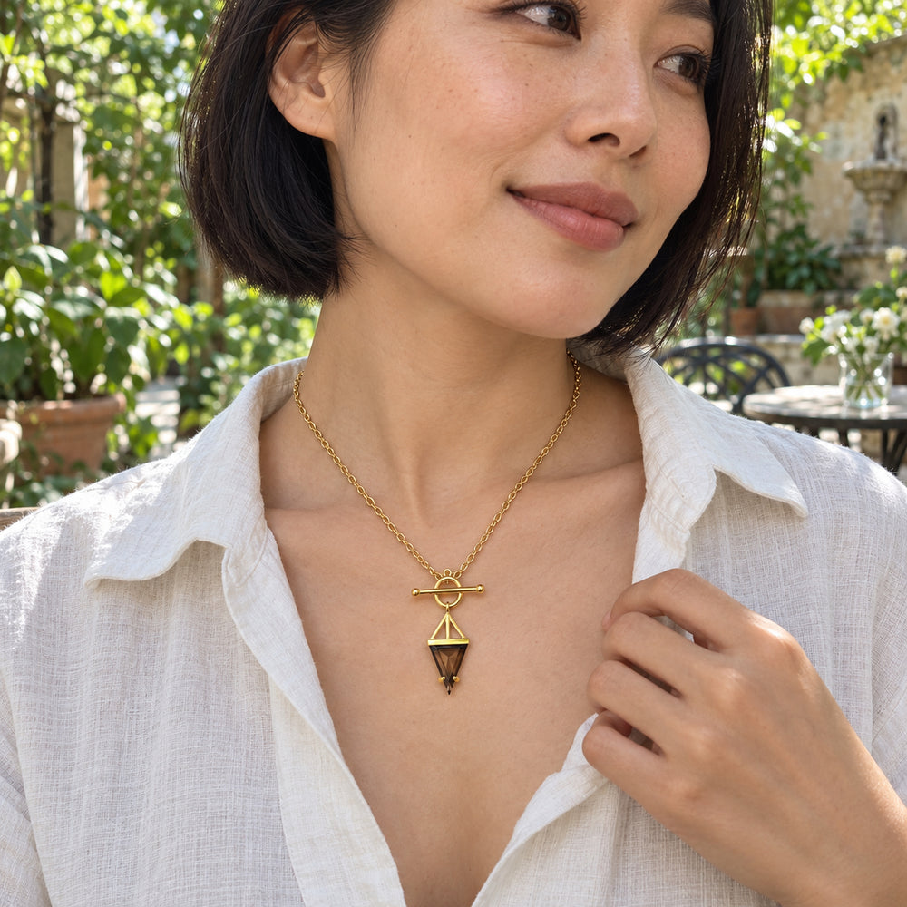 A woman with a short black bob wears the yellow gold smoky quartz necklace with a white linen blouse in a leafy courtyard cafe.